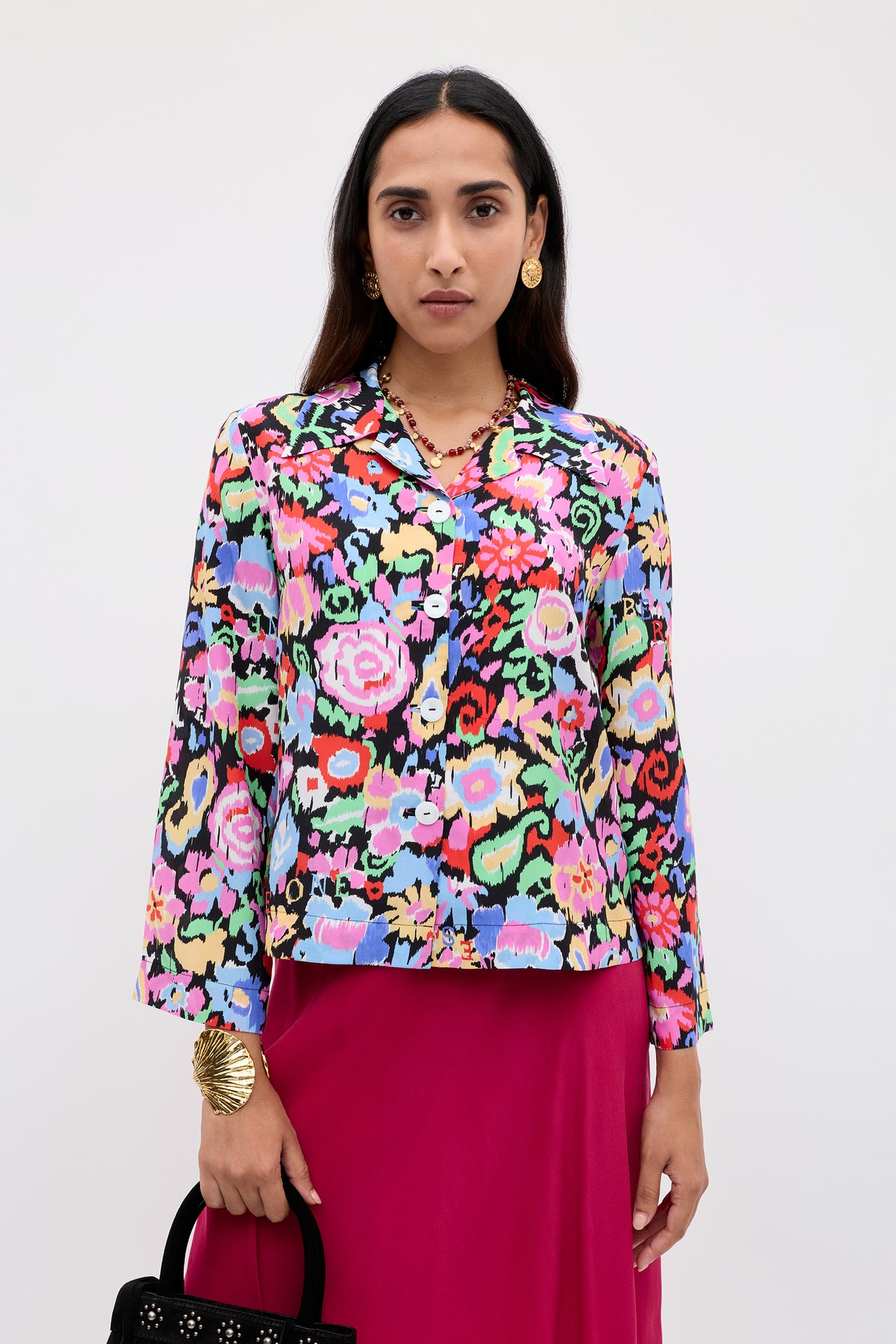A woman stands against a plain background wearing the Ginette - Ikat Bloom Charcoal long-sleeve floral blouse, a bright pink skirt, gold earrings and bracelet, and holding a black handbag.