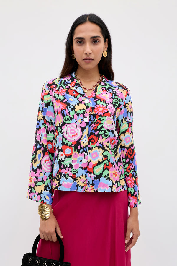 A woman stands against a plain background wearing the Ginette - Ikat Bloom Charcoal long-sleeve floral blouse, a bright pink skirt, gold earrings and bracelet, and holding a black handbag.