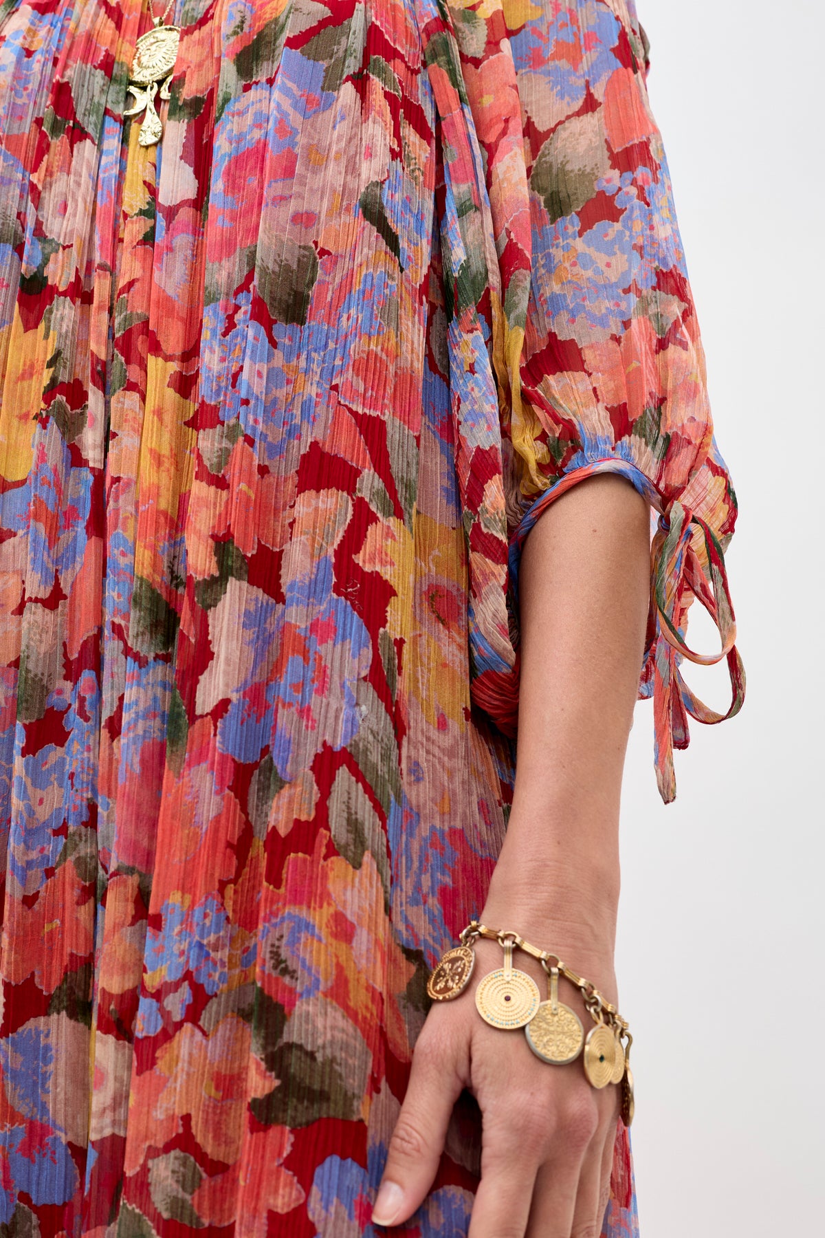 A person wearing the Hayden - Chelsea Floral Red dress with pleated fabric, along with a gold necklace and a gold charm bracelet adorned with multiple round pendants. Only one arm and the side of the dress are visible.