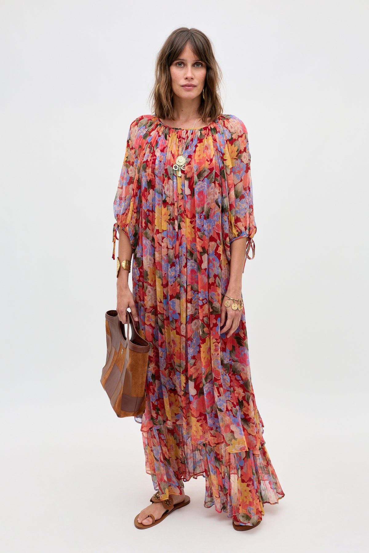 A woman wearing the Hayden - Chelsea Floral Red long, colorful floral dress with a ruffled hem, paired with brown sandals and gold jewelry, holds a brown tote bag while standing against a plain light background.