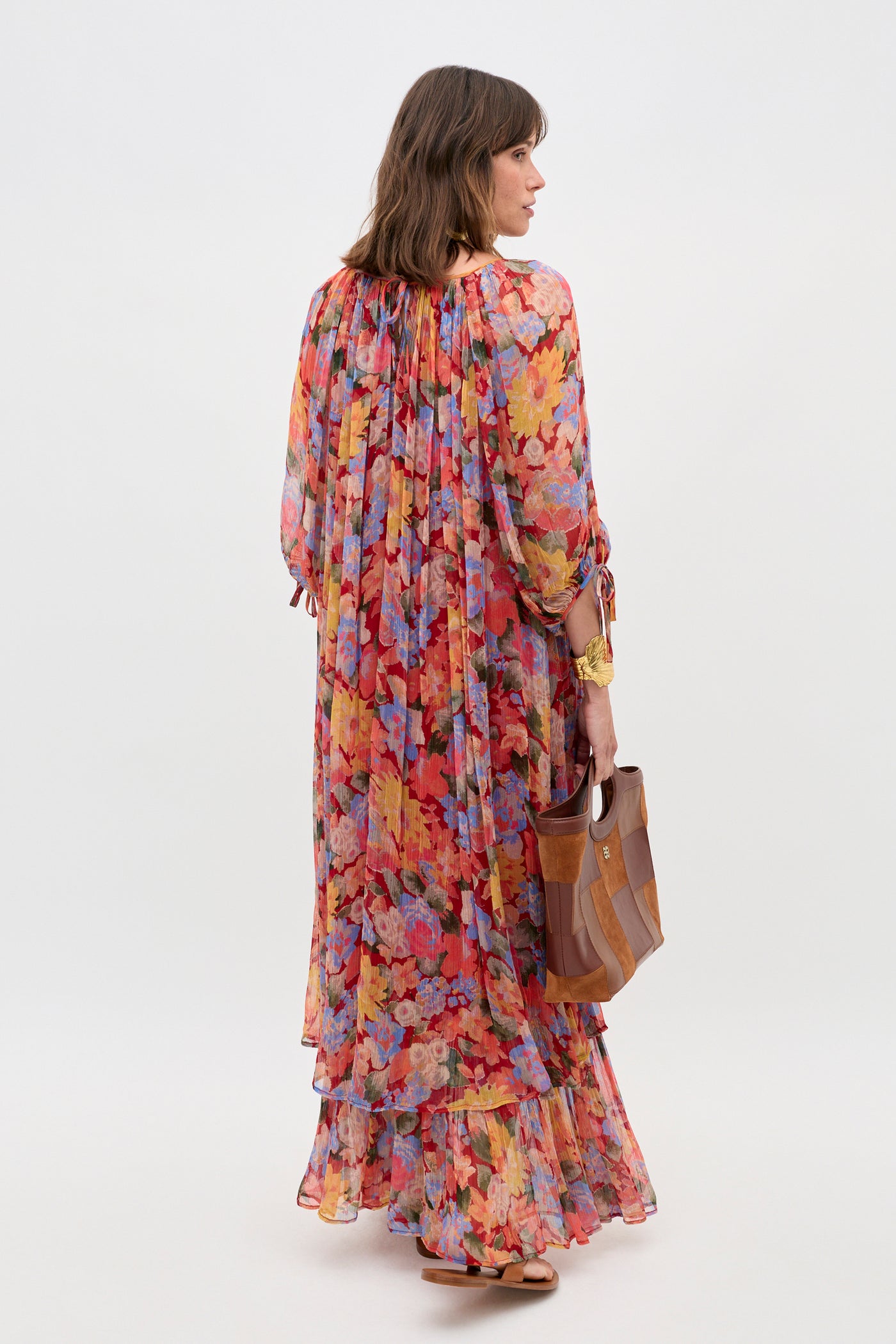 A woman stands with her back to the camera, wearing the Hayden - Chelsea Floral Red dress, which features a long, flowing layered design with vibrant floral patterns and long sleeves. She holds a brown handbag against a plain light background.
