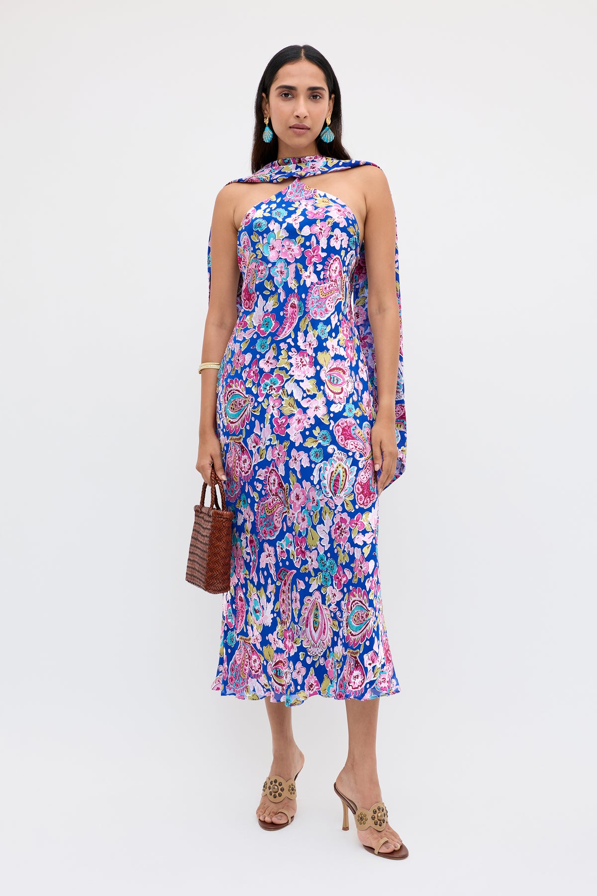 A woman wearing the Hayley - Cara Floral Devore Blue dress, featuring a colorful sleeveless design with scarf detail, holds a brown woven handbag and wears open-toed heels. She has long dark hair, statement earrings, and stands against a plain white background.