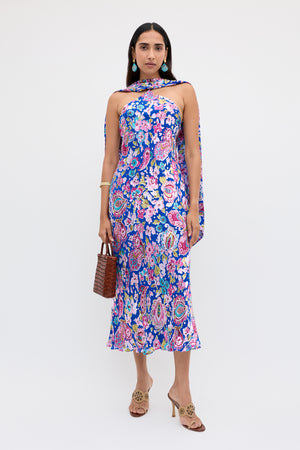 A woman wearing the Hayley - Cara Floral Devore Blue dress, featuring a colorful sleeveless design with scarf detail, holds a brown woven handbag and wears open-toed heels. She has long dark hair, statement earrings, and stands against a plain white background.