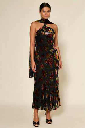 A woman wearing the Hayley - Countryside Devoré, a sleeveless, ankle-length black dress with a colorful floral pattern and halter neck, paired with open-toe black heels, stands against a plain light background.