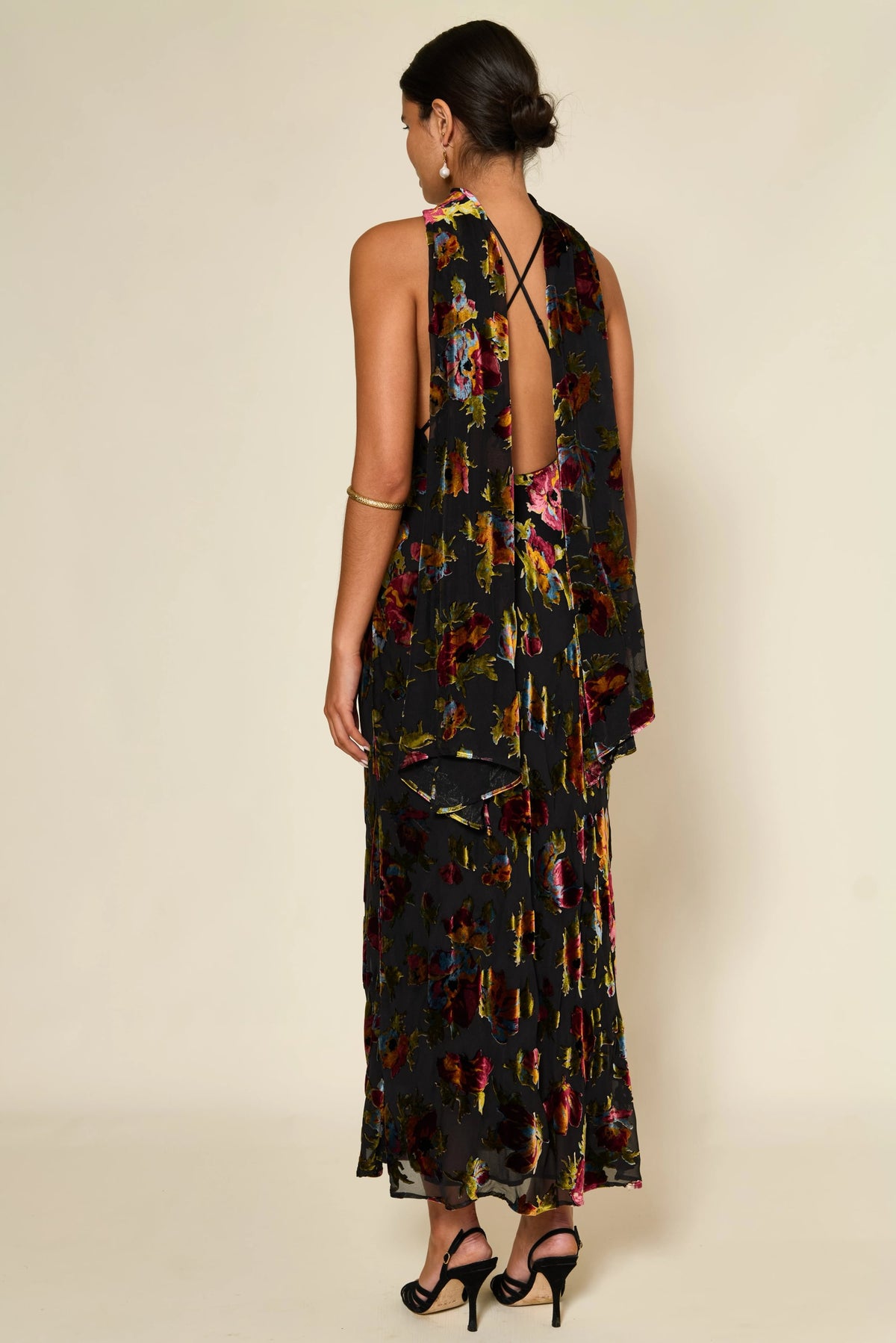 A woman stands with her back to the camera in the Hayley - Countryside Devoré, a long black floral dress featuring an open back and crossed straps. Her hair is tied back, and she accessorizes with black heels and gold jewelry.