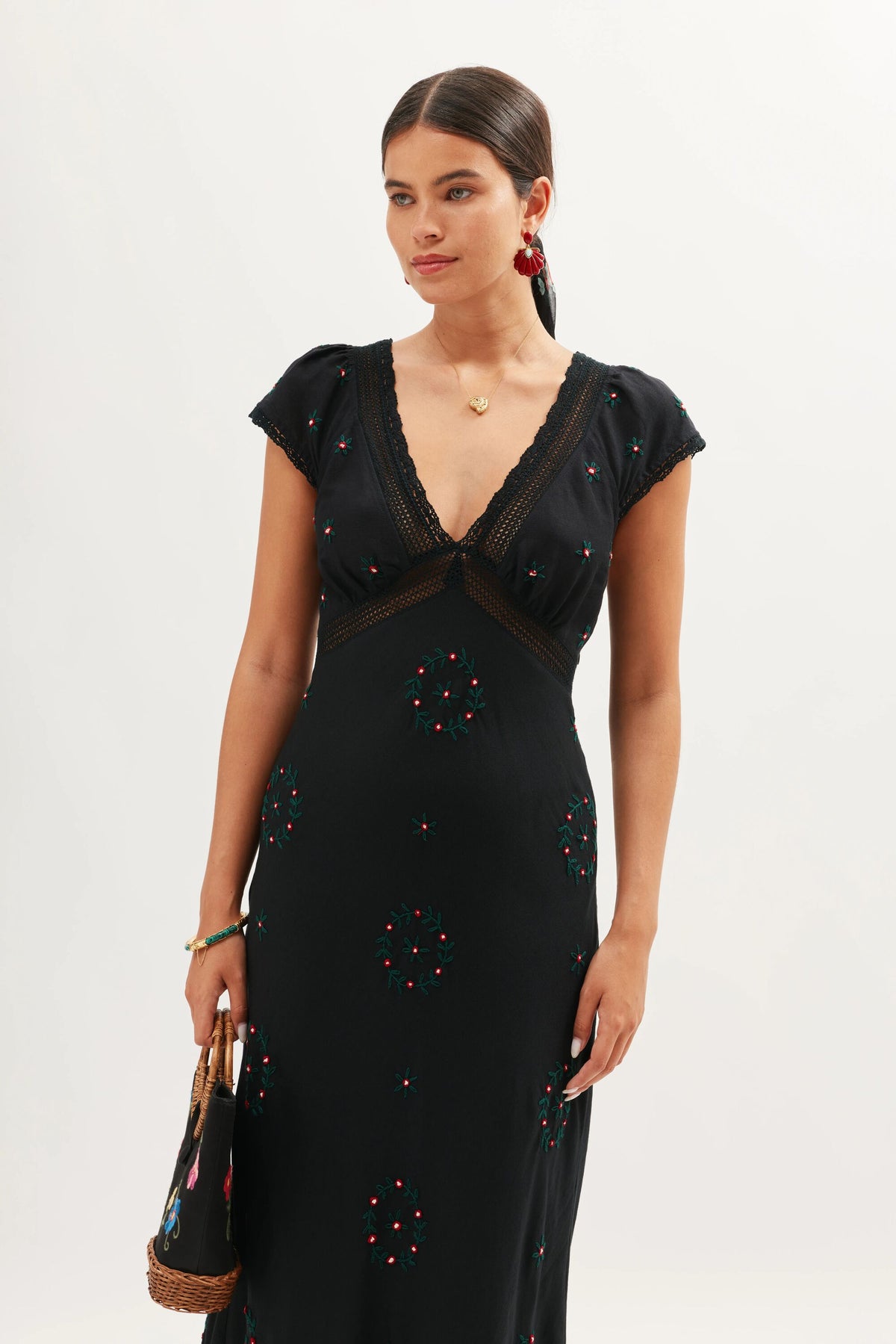 A woman with dark hair in a low ponytail wears the Iggy - Rose Trail Embroidery Black dress, showcasing its deep V-neckline and floral embroidery. She holds a woven handbag and finishes the look with red earrings and a gold necklace.