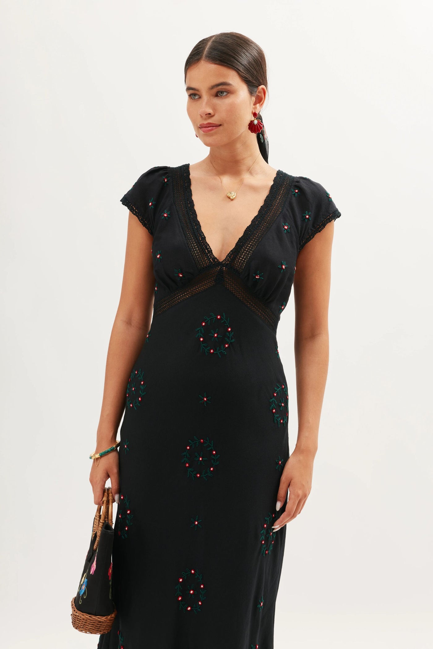 A woman with dark hair in a low ponytail wears the Iggy - Rose Trail Embroidery Black dress, showcasing its deep V-neckline and floral embroidery. She holds a woven handbag and finishes the look with red earrings and a gold necklace.