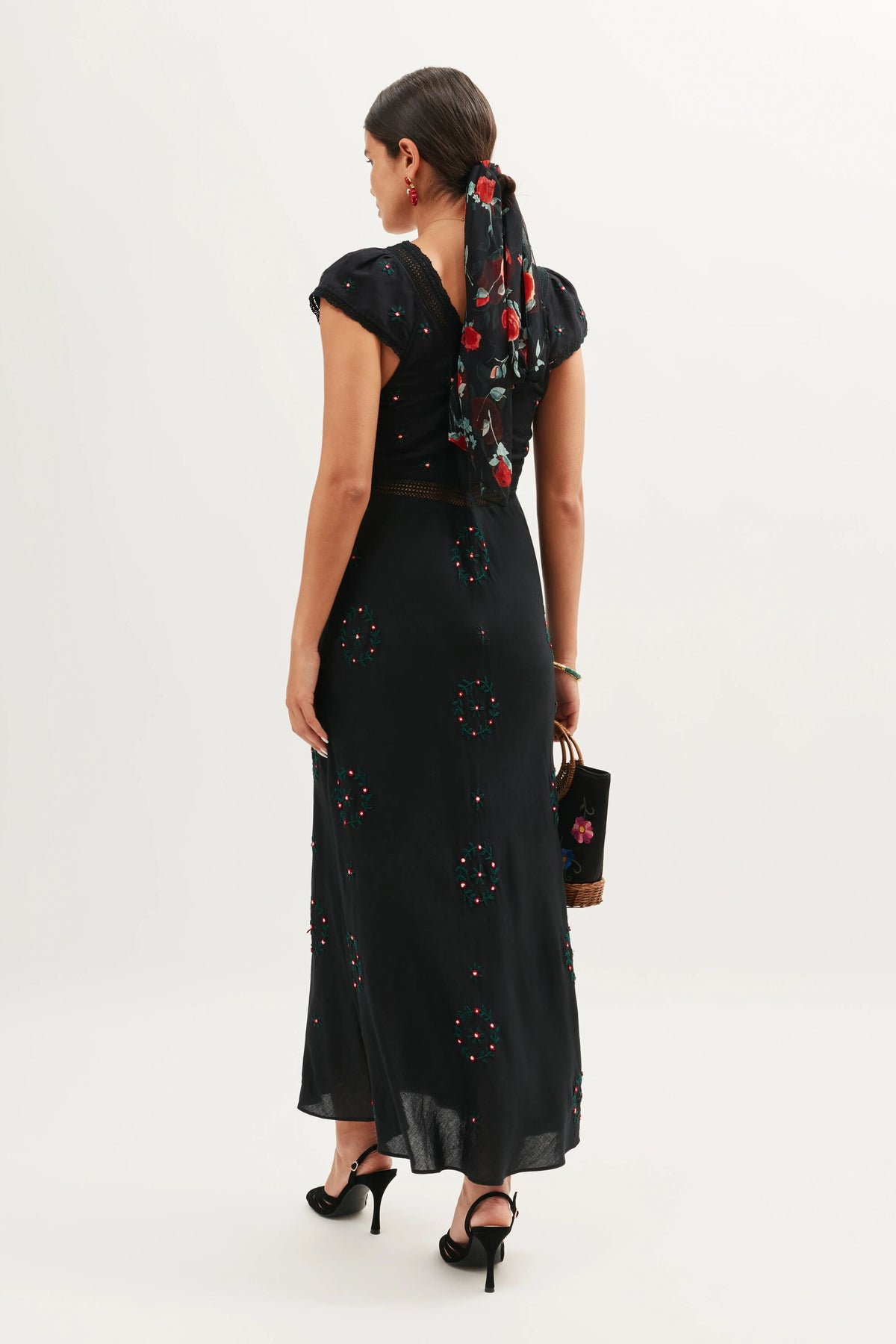 A woman faces away, wearing the Iggy - Rose Trail Embroidery Black dress—an ankle-length black dress with colorful floral embroidery. She pairs it with a floral scarf in her hair, black heels, and a black flower-designed handbag.