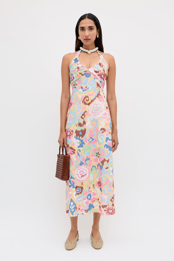 A woman with long, straight hair and a calm expression stands against a white background wearing the Ilyana - Ikat Bloom Yellow halter dress, beige woven shoes, a shell necklace, and holding a small brown woven handbag.