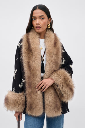 A woman wearing the Micah - Alexa Flower Embroidery Black jacket with light brown faux fur trim on the collar and cuffs, paired with blue jeans, gold earrings, and layered necklaces, stands against a plain light background.