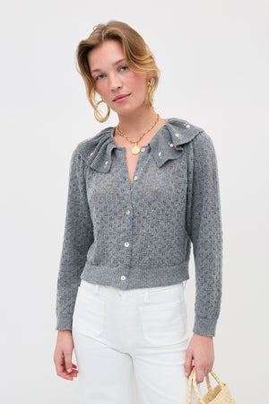 A woman wearing the Jelena - Luna Bud gray pointelle-knit button-up sweater with a ruffled collar and pearl details, styled with high-waisted white pants and a woven bag, stands against a plain white background.
