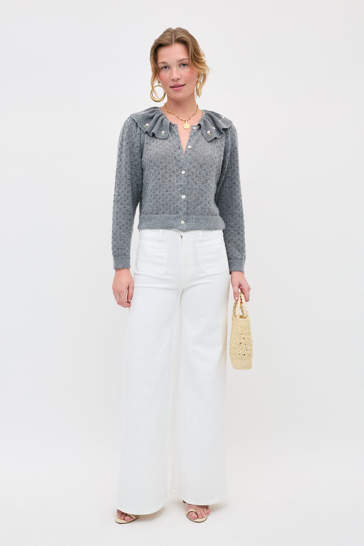 A woman stands against a white background wearing the Jelena - Luna Bud gray pointelle-knit cardigan with a large collar, paired with white wide-leg pants, sandals, and holding a small woven handbag.