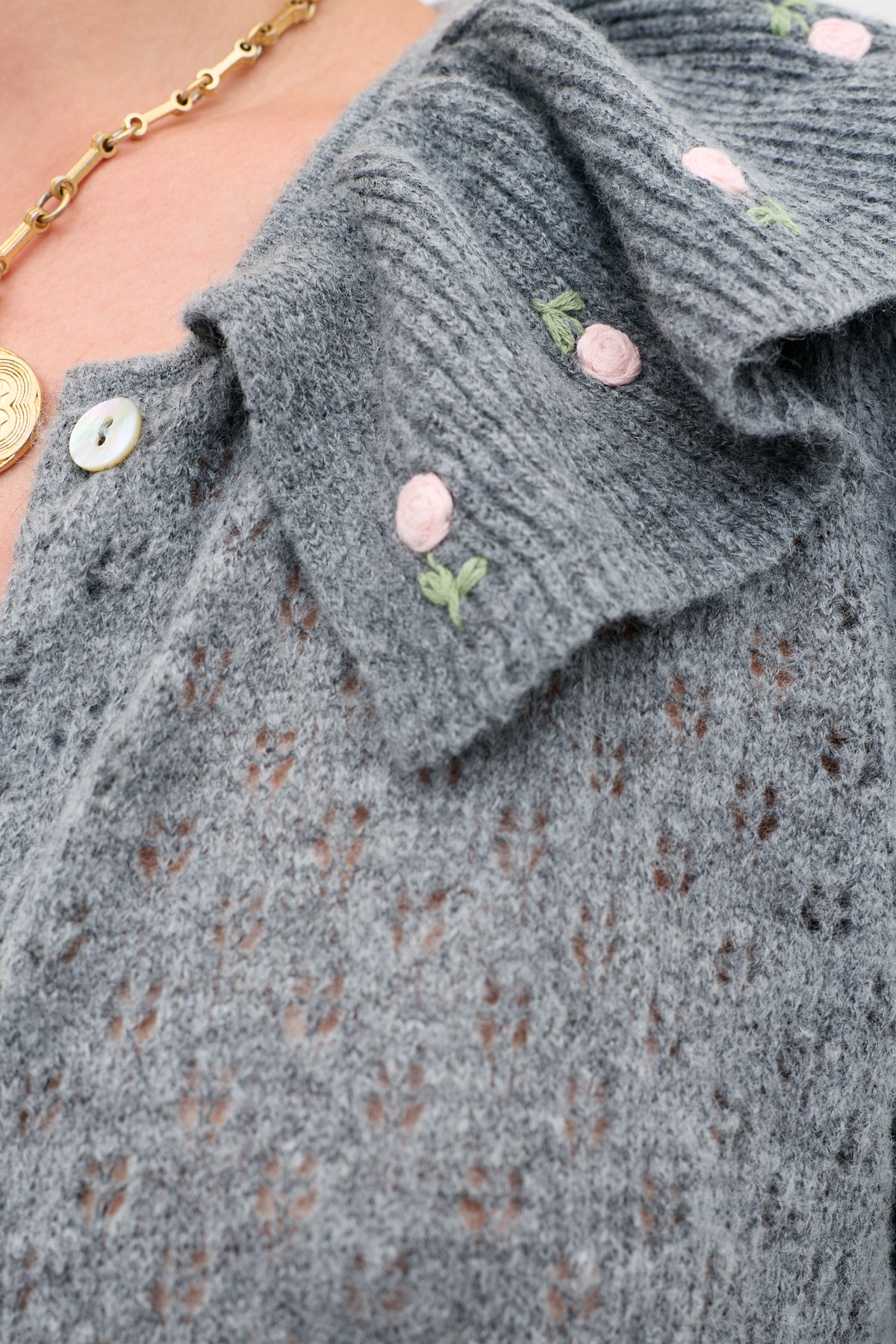 Close-up of the Jelena - Luna Bud gray pointelle-knit sweater, featuring delicate pink floral embroidery. A gold chain necklace and a white button are visible near the collar.