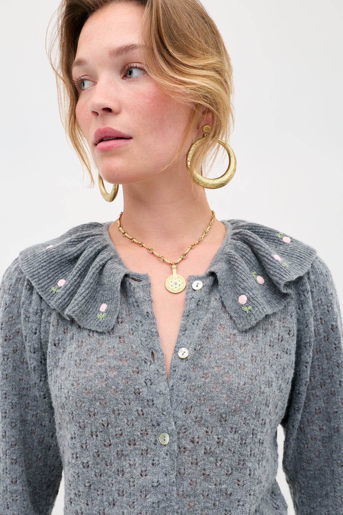A woman wears the Jelena - Luna Bud gray pointelle-knit cardigan featuring a ruffled, floral-embroidered collar, paired with large gold hoop earrings and a gold chain necklace with a coin pendant, as she gazes slightly to the side against a plain background.
