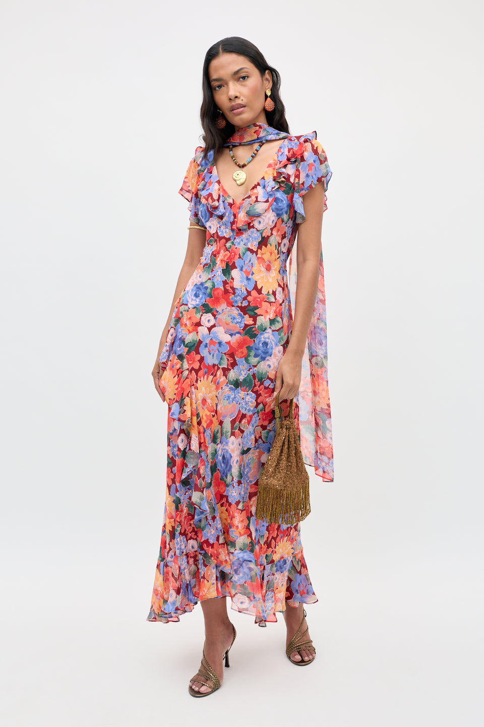 A woman stands against a plain white background wearing the Julien - Chelsea Floral Red dress, styled with ruffles. She accessorizes with a matching floral scarf, gold jewelry, brown fringed handbag, and heeled sandals.