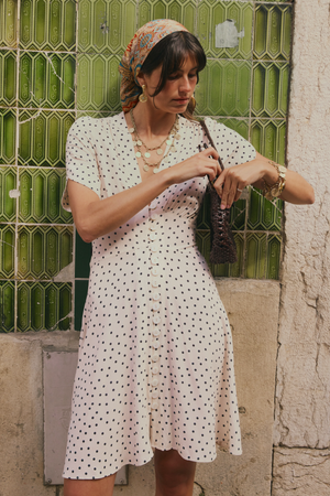 A woman wearing the Sakura - Layla Dot Ivory dress stands before a textured green glass wall, adjusting a small woven bag on her shoulder. She accessorizes with a colorful headscarf, layered necklaces, and hoop earrings.