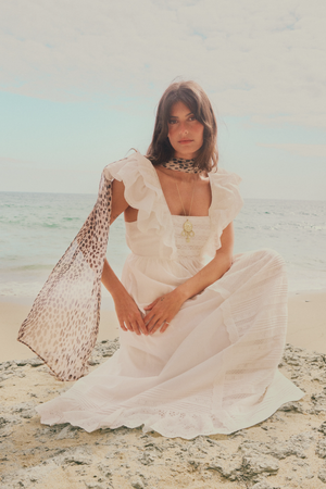 A woman wearing the Delaney - White dress kneels on a sandy beach with the ocean and sky in the background. She has long brown hair, a leopard print scarf, and layered necklaces.