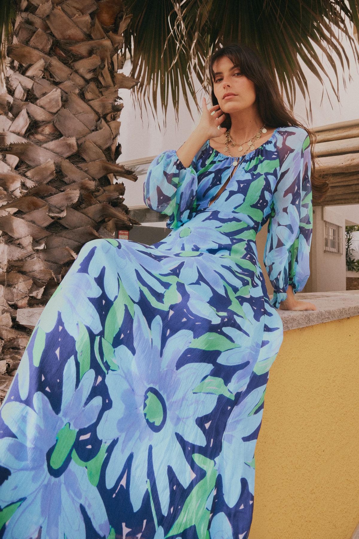 A woman in the Nikolette - Danish Floral Blue maxi dress sits on a yellow wall by a palm tree, looking thoughtful with her hand near her face.