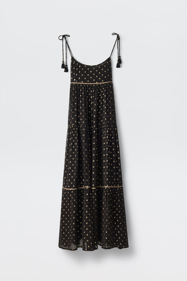 The Kamilah - Black is a sleeveless maxi dress featuring thin shoulder straps with tassel details, decorated with small light-colored polka dots and tiered layers, shown hanging against a plain white background.