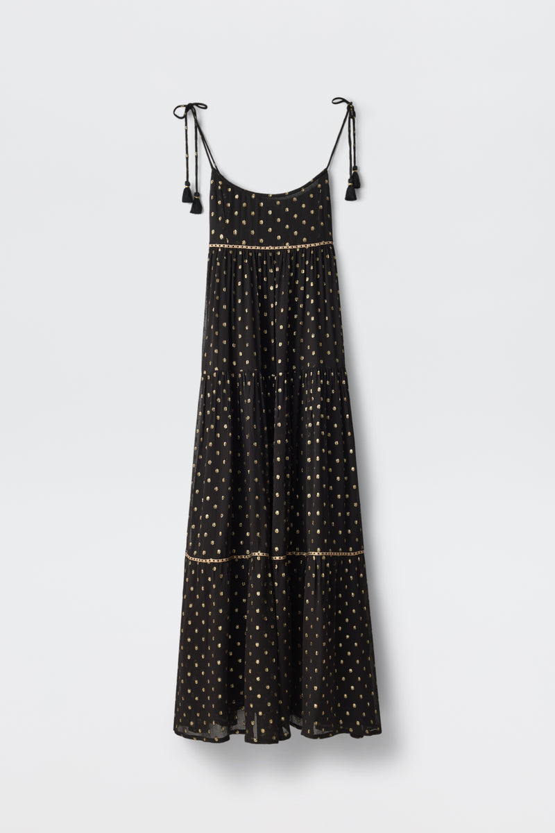 The Kamilah - Black is a sleeveless maxi dress featuring thin shoulder straps with tassel details, decorated with small light-colored polka dots and tiered layers, shown hanging against a plain white background.