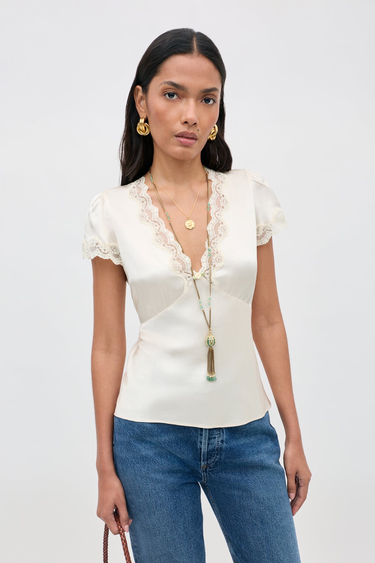 A woman wearing the Karia - Champagne cream satin short-sleeve blouse with lace trim, paired with blue jeans, gold earrings, and layered gold necklaces, stands against a plain light background.