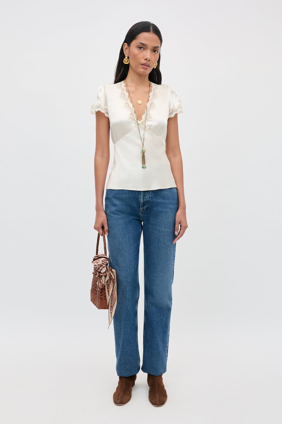 Wearing the Karia - Champagne blouse, a woman pairs it with blue jeans, brown ankle boots, and carries a brown woven handbag. She accessorizes with gold jewelry and a long necklace, standing against a plain light-colored background.