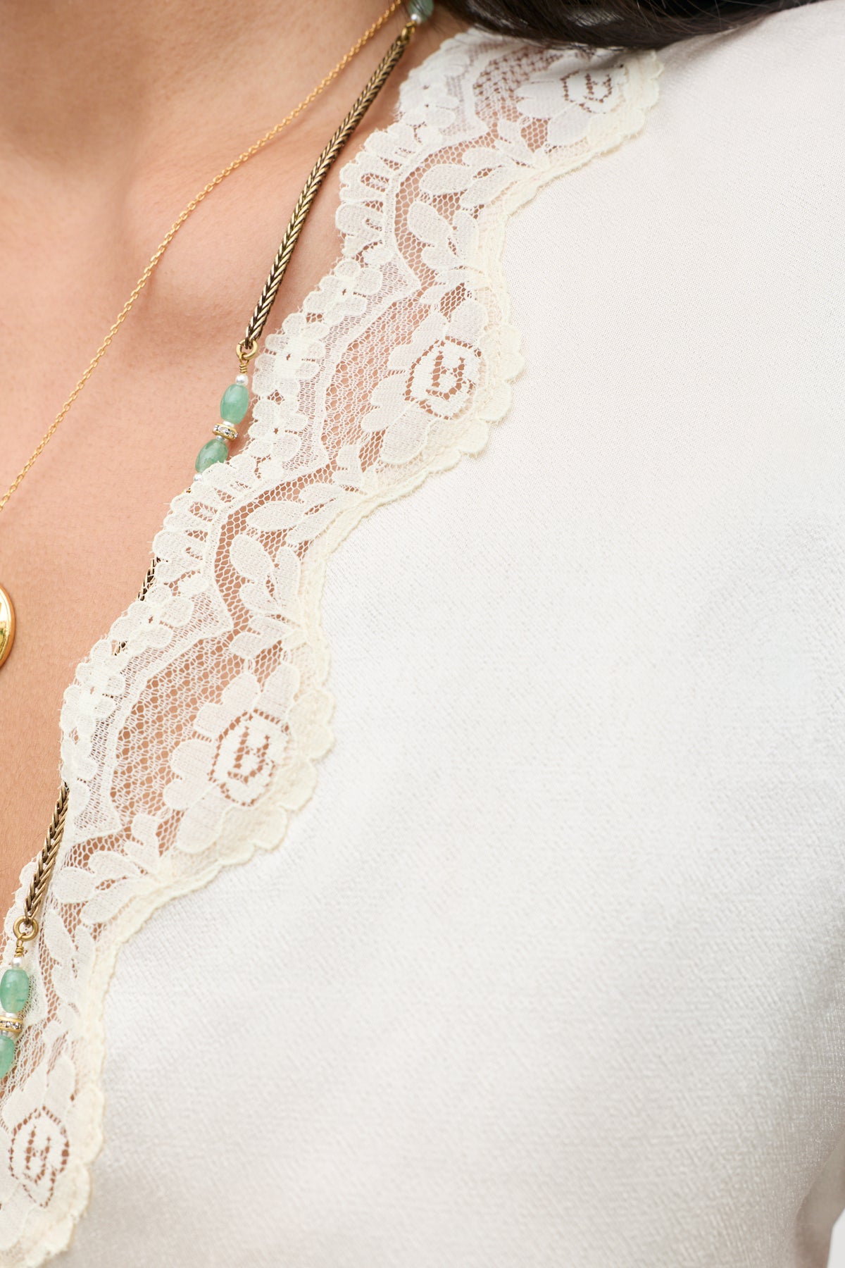Close-up of a woman’s neckline wearing the Karia - Champagne top featuring a scalloped lace trim, styled with layered gold necklaces adorned with turquoise beads.