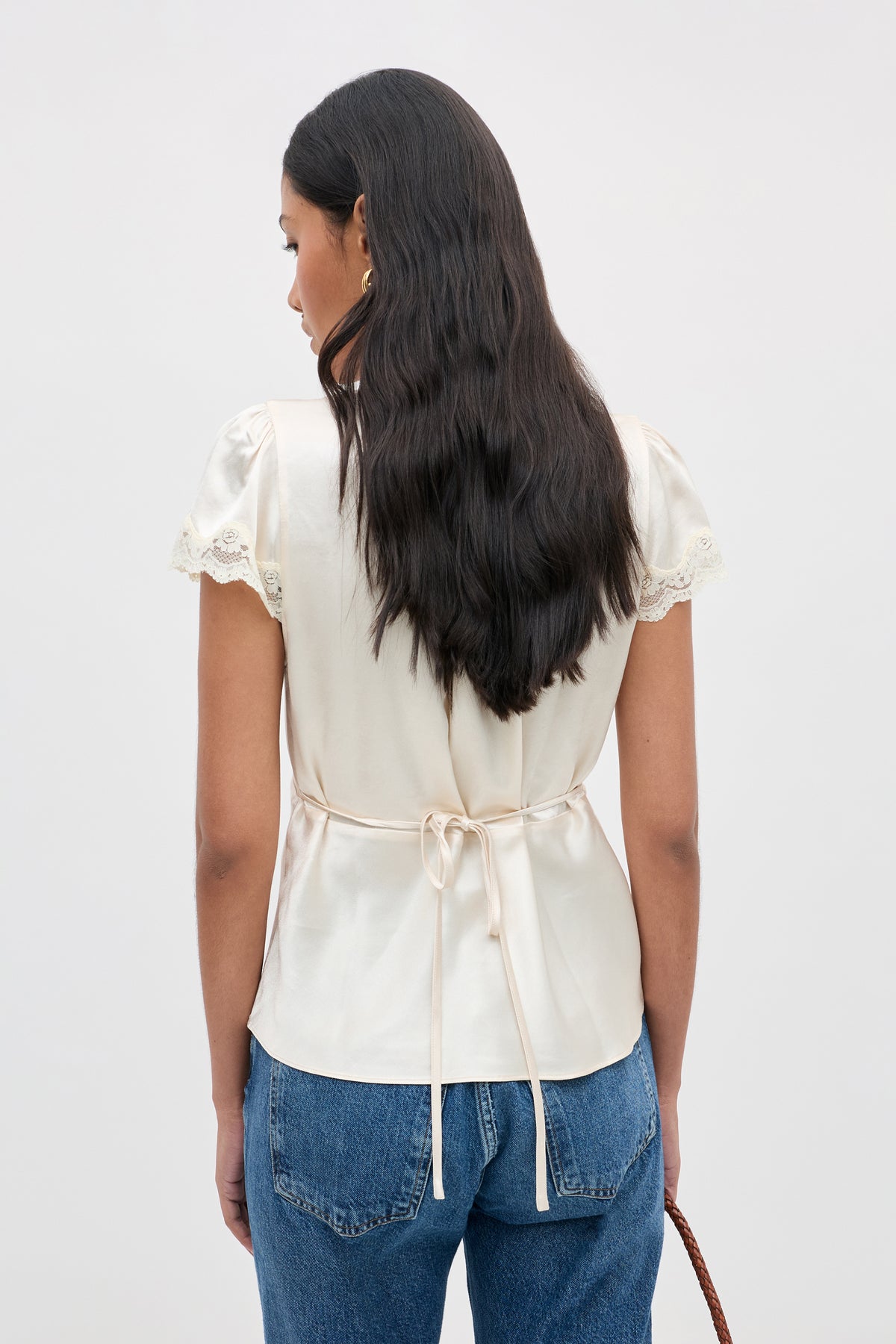 A woman stands with her back to the camera, wearing the Karia - Champagne satin blouse featuring lace-trimmed short sleeves. She pairs it with blue jeans and holds a brown bag, her long, dark hair loose and wavy.