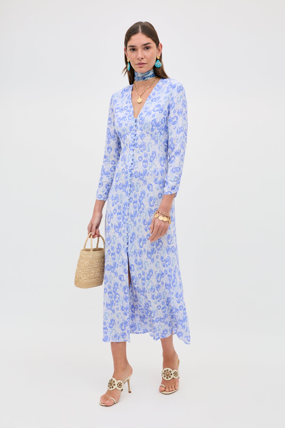 A woman wearing the Katie - Tulip Fields Cornflower Blue dress with beige heeled sandals, turquoise earrings, layered necklaces, a patterned neck scarf, and a straw handbag stands against a white background—perfect for spring and summer.