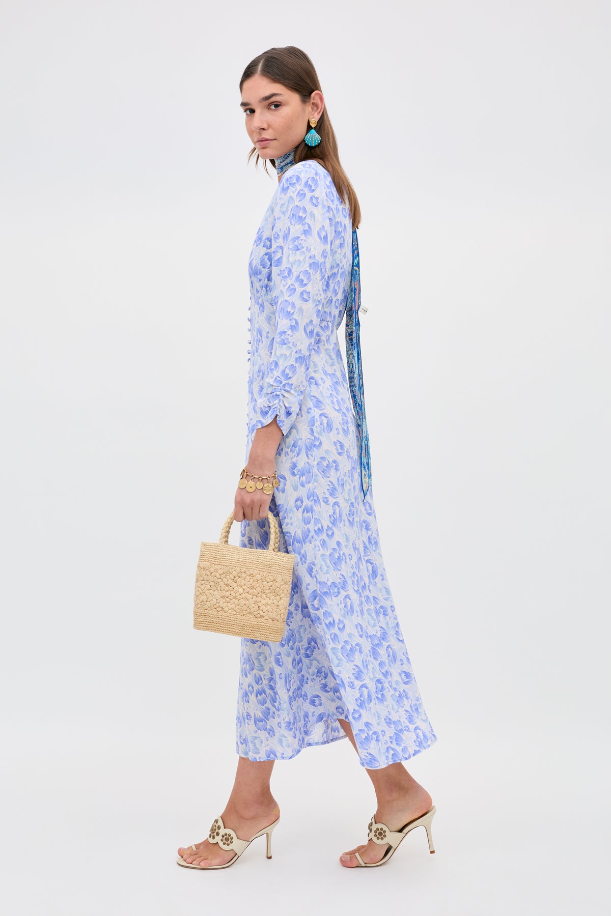 A woman in the Katie - Tulip Fields Cornflower Blue dress, beige heeled sandals, and turquoise earrings holds a small woven straw handbag. Perfect for spring and summer, she poses against a plain white background, looking over her shoulder.