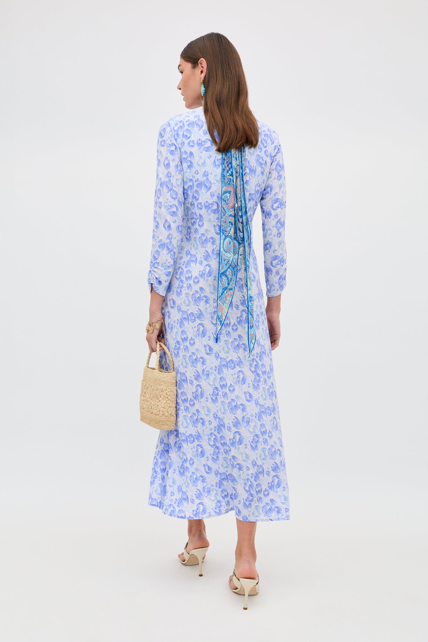 A woman stands facing away, wearing the Katie - Tulip Fields Cornflower Blue dress—ideal for spring and summer. She accessorizes with a woven handbag, patterned hair scarf, white heeled sandals, and statement earrings.