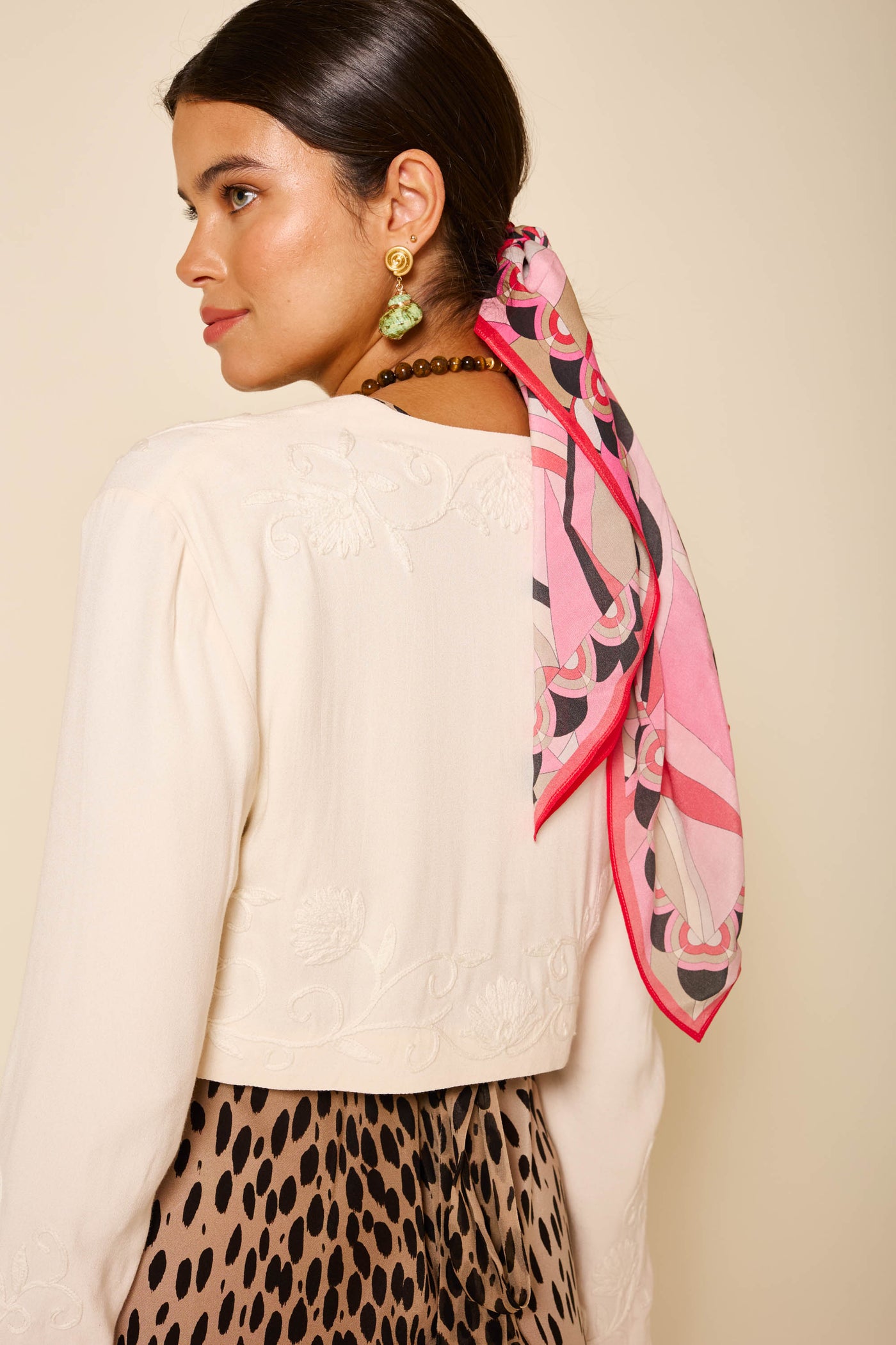 A woman with dark hair tied back wears the Learke - Cream cropped vintage jacket with floral embroidery, a pink patterned scarf, and a leopard-print skirt, facing away but glancing over her shoulder.