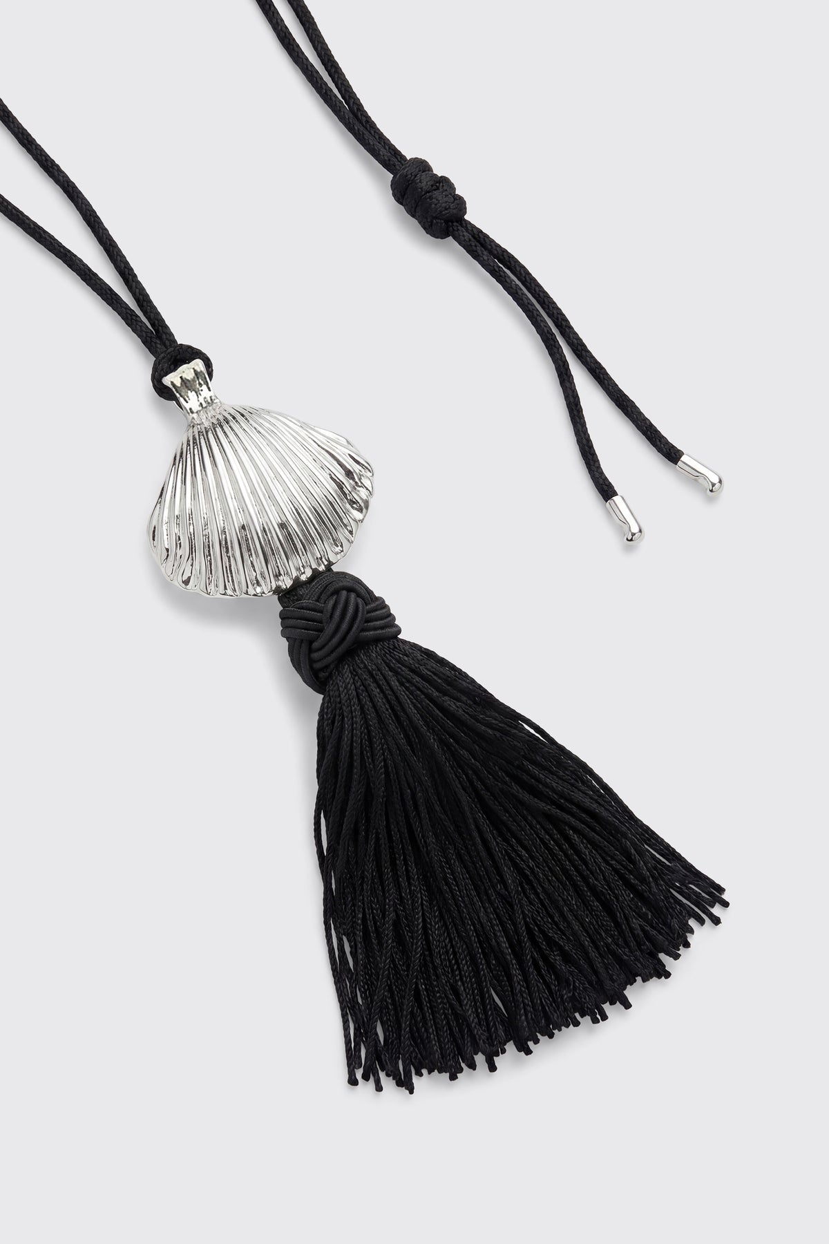 Liana - Silver: A necklace featuring a silver shell pendant and a black knotted tassel, suspended from a black cord with silver tips, displayed on a light gray background.