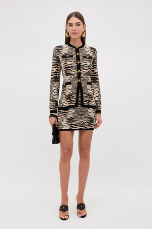 A woman stands facing forward against a plain white background, wearing the Liesel - Cooper Tiger Beige set—a tiger-striped jacket and matching mini skirt—paired with black heeled sandals, gold earrings, and holding a small black clutch.
