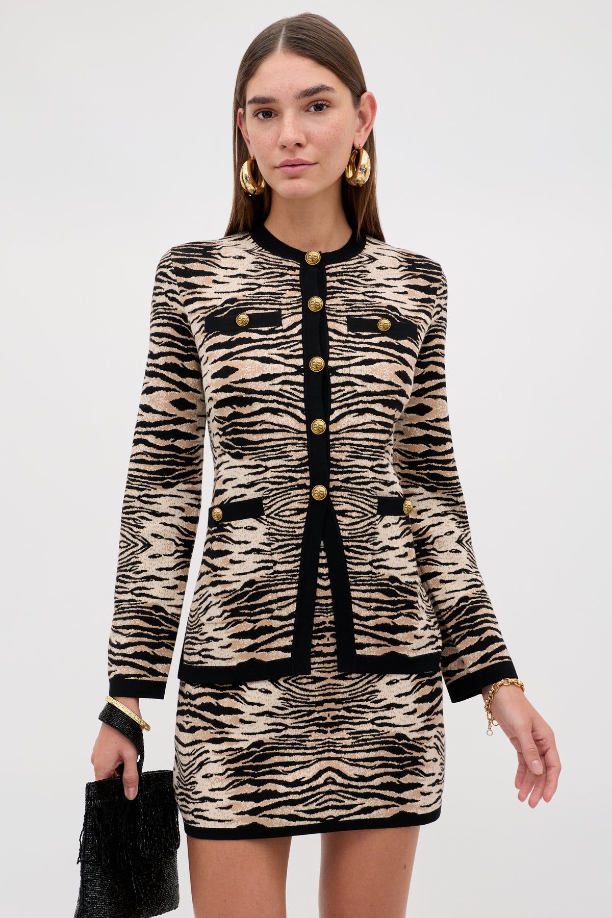 Wearing the “Liesel - Cooper Tiger Beige” set featuring a beige and black animal print jacket and skirt with gold buttons, she completes her look with gold hoop earrings and a black textured handbag while standing against a plain light background.