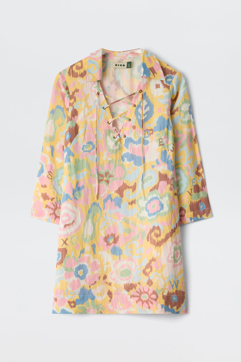 The Lucien - Ikat Bloom Yellow is a long-sleeve shirt with a pastel floral print in pink, yellow, blue, and green, inspired by Ikat Bloom. It features a lace-up V-neck and hidden letters scattered within the design.