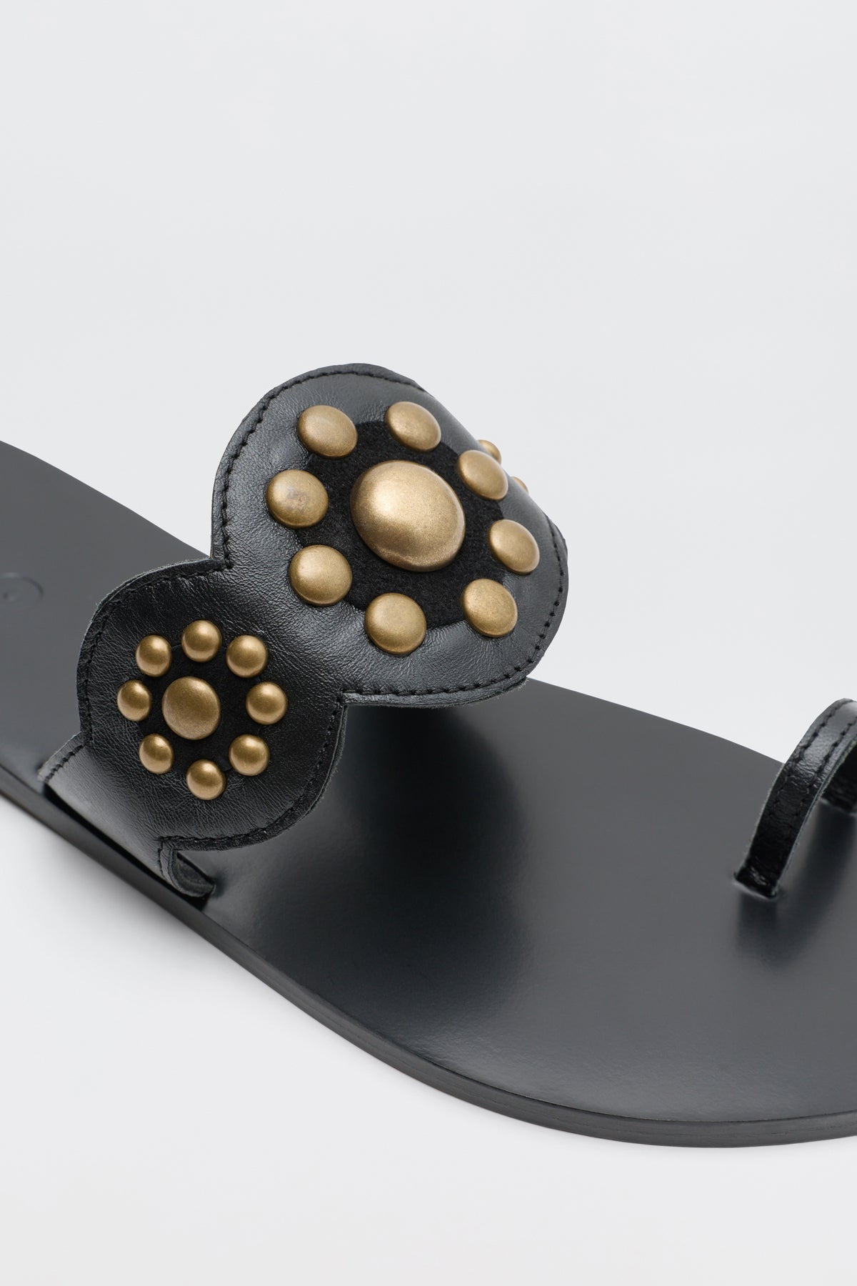Lucille - Black sandal features a wide strap adorned with gold circular studs in a flower-like arrangement, complemented by a thin toe loop. The sandal is displayed on a plain white background.