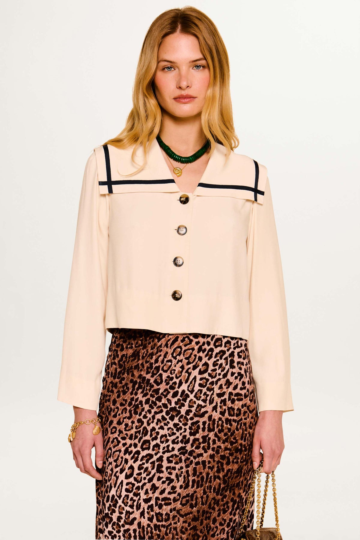 A woman with long blonde hair wears the Manon - Almond jacket with black trim and a leopard print skirt, standing against a plain white background and holding a gold chain handbag.