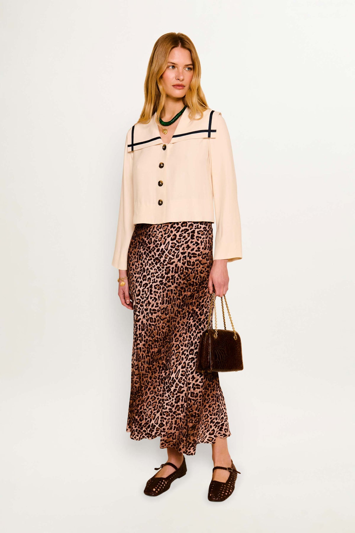 A blonde woman stands against a white background wearing the Manon - Almond jacket with black trim, a leopard print skirt, brown shoes, and holding a brown handbag. She accessorizes with a green necklace for a modern nautical look.