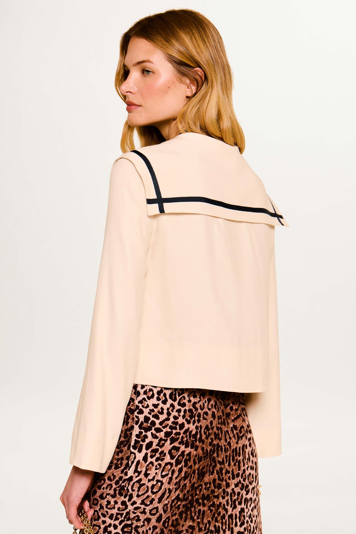 A woman with medium-length blonde hair wears the Manon - Almond jacket with dark trim and a leopard print skirt, standing against a plain white background, facing away and slightly to the side.