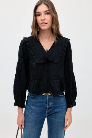 Woman with wavy brown hair wears the Maribelle - Black, a vintage-inspired blouse featuring long sleeves, ruffled details, a large collar, and a relaxed fit. She pairs it with flattering blue jeans and holds a small brown purse while standing against a plain light background.