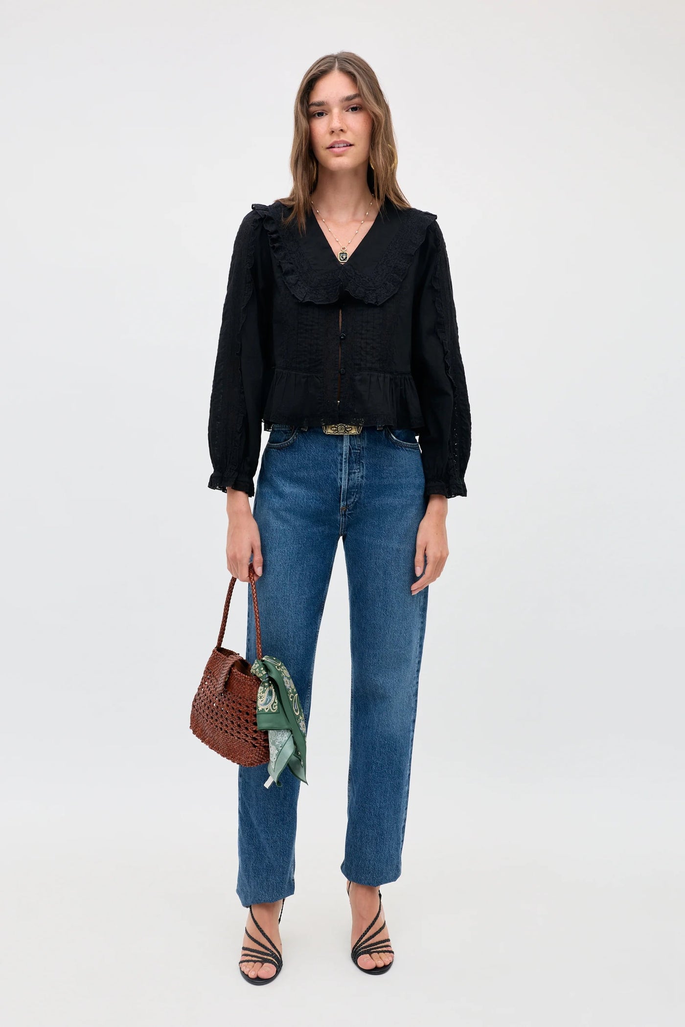 Woman stands against a plain background, wearing the Maribelle - Black long-sleeve blouse with a relaxed fit, paired with blue jeans and black strappy sandals. She holds a brown woven handbag with a green scarf tied to it.
