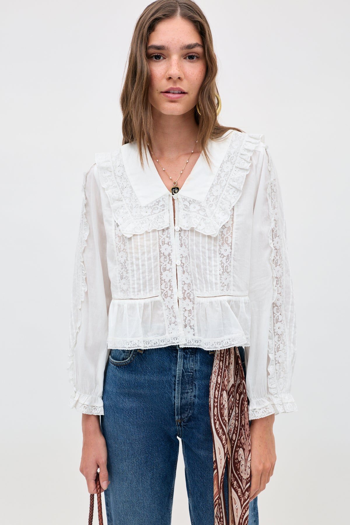 A woman wears the Maribelle - Ivory, a vintage-inspired white blouse with lace accents and a ruffled collar, paired with blue jeans. She stands against a plain background, holding a bag adorned with a patterned scarf.