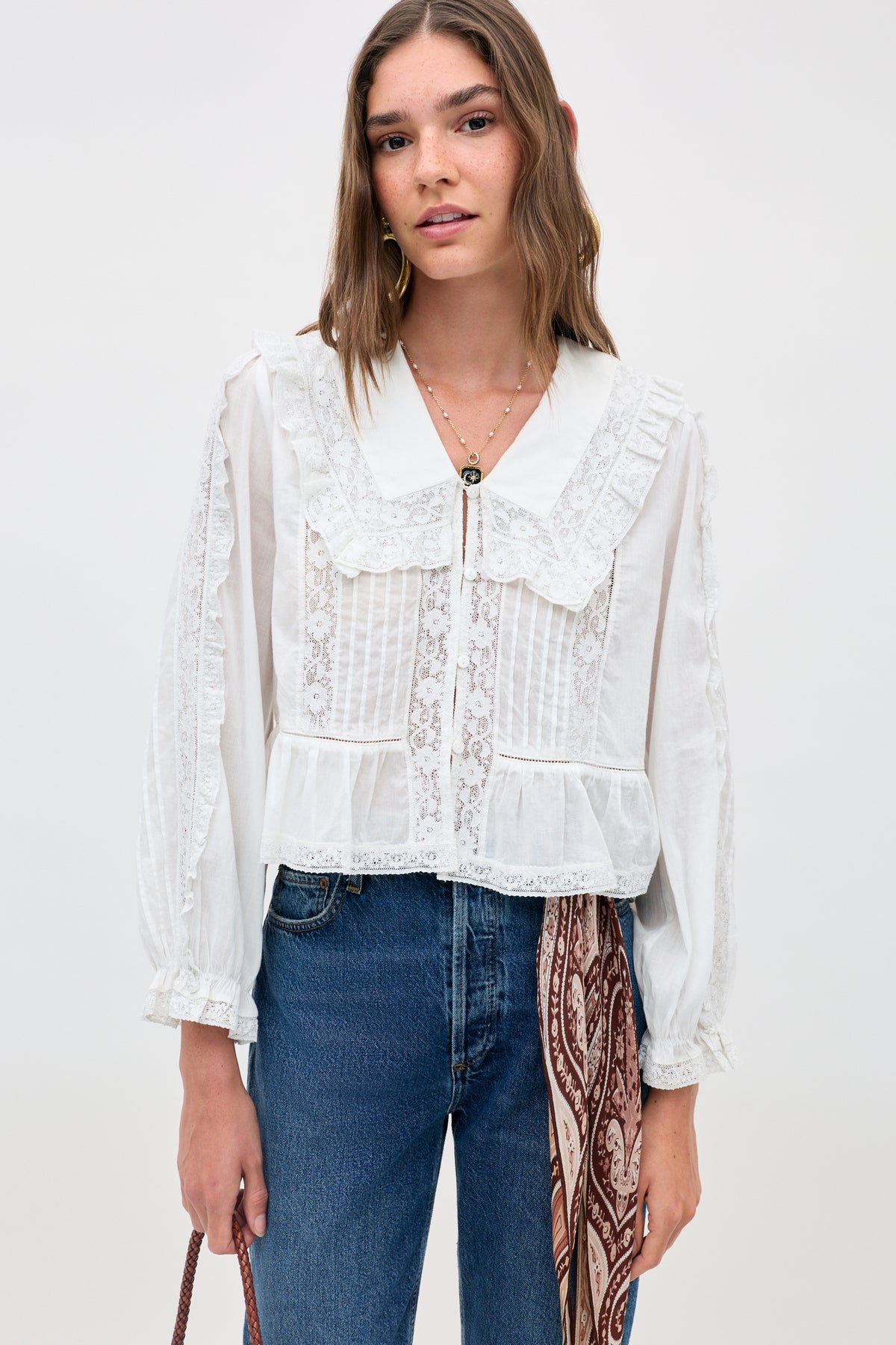 Woman wearing the Maribelle - Ivory blouse, a white long-sleeved top with lace details and a ruffled collar, styled with blue jeans and a brown patterned scarf at the waist, stands against a plain light background.