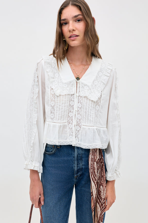 Woman wearing the Maribelle - Ivory blouse, a white long-sleeved top with lace details and a ruffled collar, styled with blue jeans and a brown patterned scarf at the waist, stands against a plain light background.