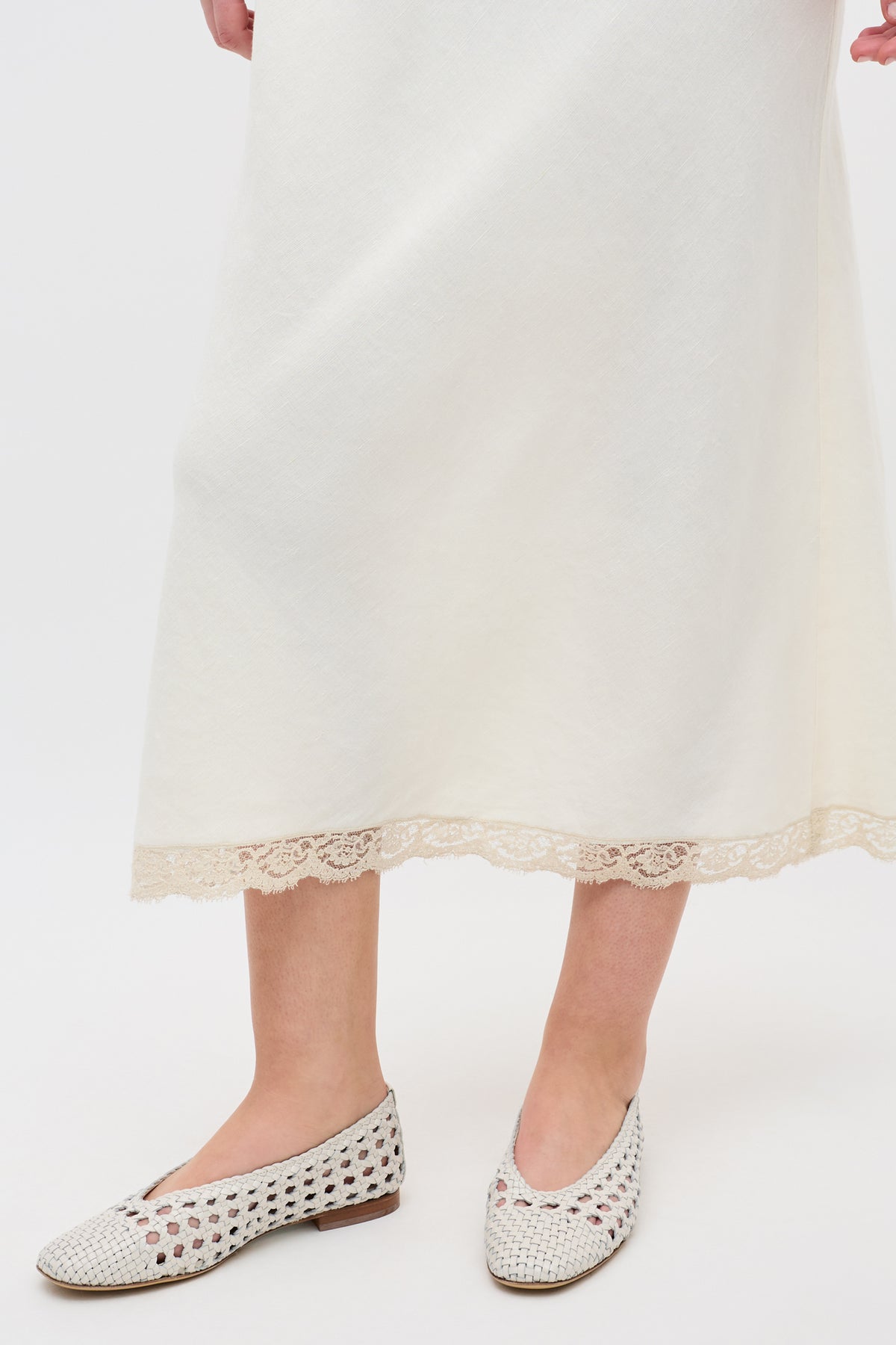 Someone is wearing the Marlia - Cream, a long off-white skirt with lace-trimmed hem, paired with white woven slip-on shoes and posing against a plain white background.