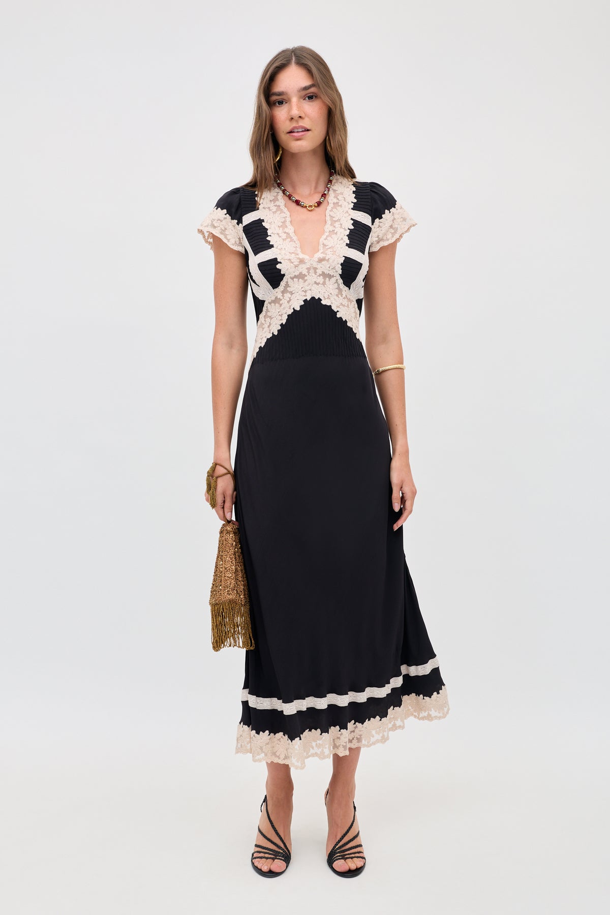 A woman poses against a plain backdrop in the Martina - Black midi dress with cream lace trim, short sleeves, and a deep V-neck. She accessorizes with black strappy heels, a gold fringed bag, necklace, and bracelet.
