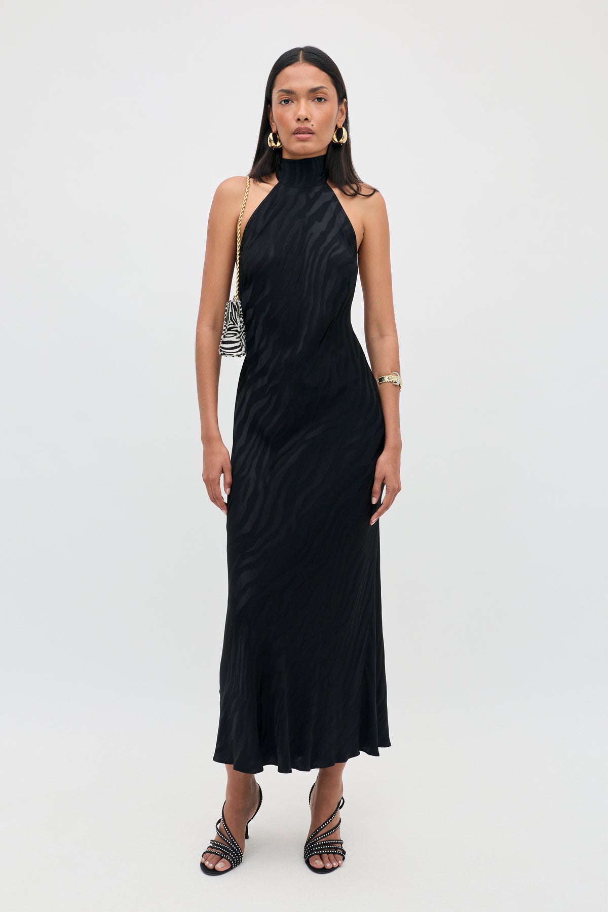 A woman stands against a plain white background wearing the Maura - Cooper Jacquard Black dress, a sleeveless, high-neck, ankle-length black piece, paired with strappy black heels, gold earrings, a gold bracelet, and a small zebra-print shoulder bag.