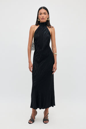 A woman stands against a plain white background wearing the Maura - Cooper Jacquard Black dress, a sleeveless, high-neck, ankle-length black piece, paired with strappy black heels, gold earrings, a gold bracelet, and a small zebra-print shoulder bag.