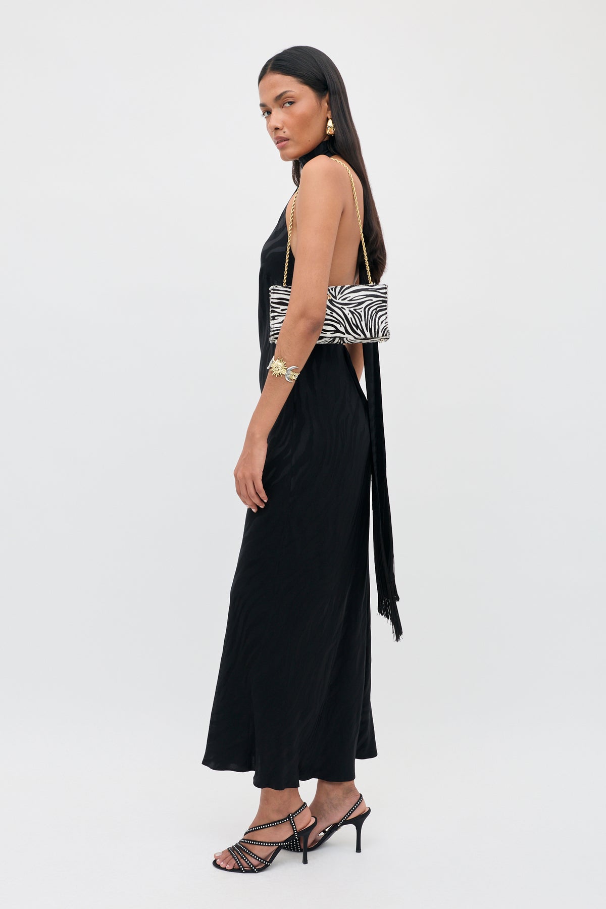 A woman in the Maura - Cooper Jacquard Black sleeveless dress and heeled sandals stands in profile, holding a zebra-print purse with a gold chain strap. She has long dark hair and accessorizes with gold statement earrings and a bracelet.